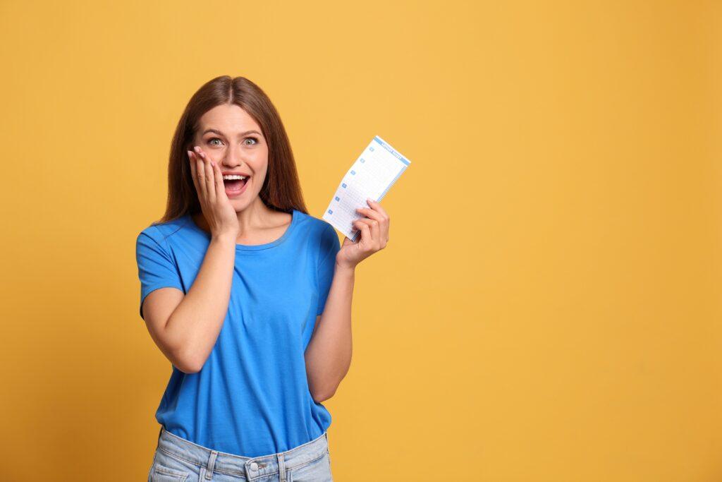 Eine glückliche Frau im blauen T-Shirt hält einen Lottoschein und zeigt eine freudige Reaktion vor einem gelben Hintergrund.