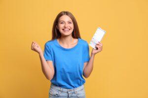 Eine lachende Frau im blauen T-Shirt hält einen Lottoschein und zeigt eine jubelnde Geste vor einem gelben Hintergrund.