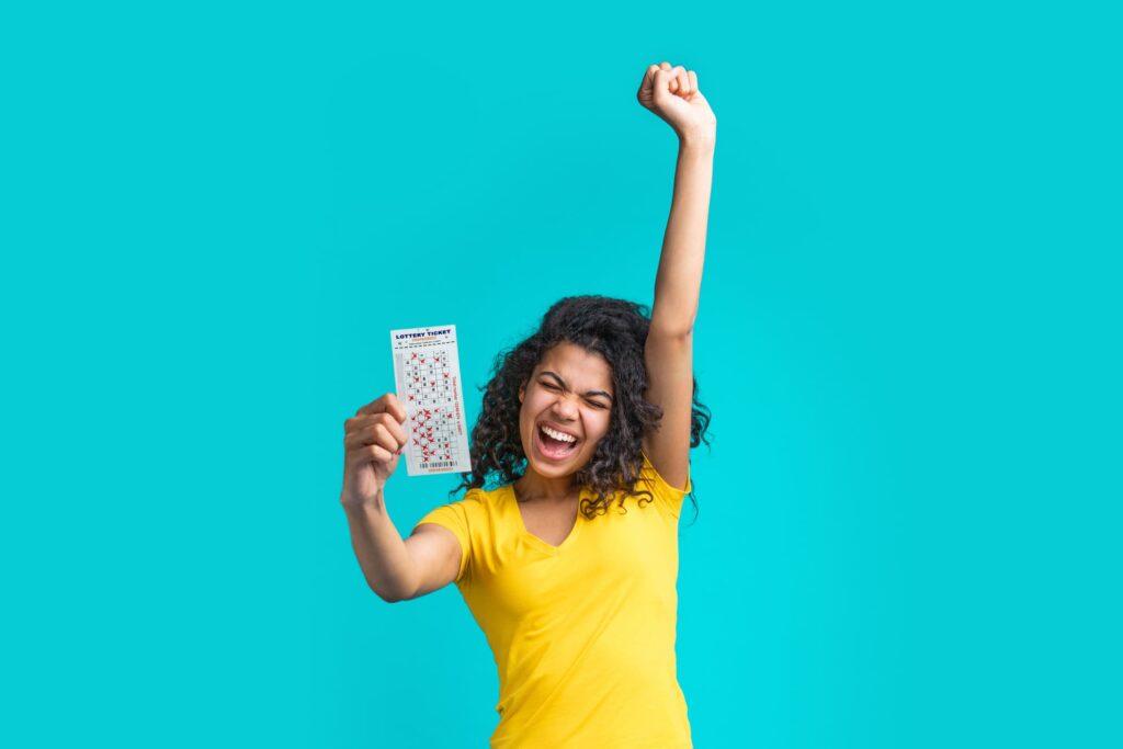Eine strahlende Frau in einem gelben T-Shirt jubelt und hält stolz ihren Lottoschein hoch. Die Freude über das Eurojackpot online spielen ist deutlich sichtbar, während sie vor einem hellblauen Hintergrund posiert.