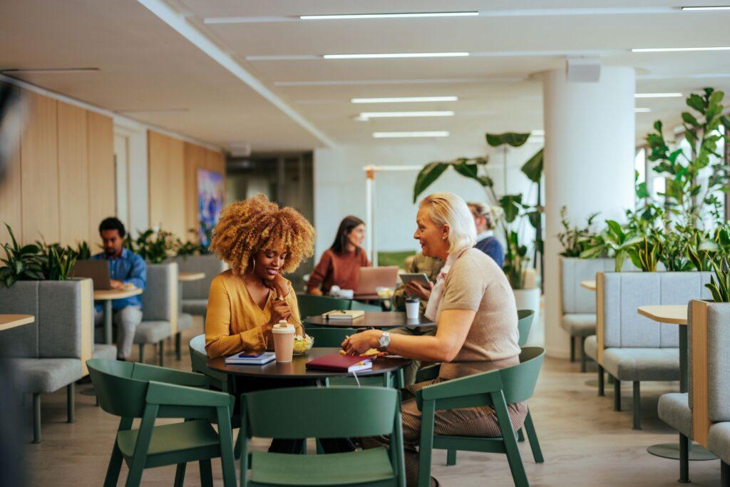 Mitarbeiter genießen eine gesunde Mahlzeit in einem modernen Betriebsrestaurant. Die entspannte Atmosphäre und das grüne, pflanzenreiche Umfeld fördern den Austausch und das Wohlbefinden am Arbeitsplatz.