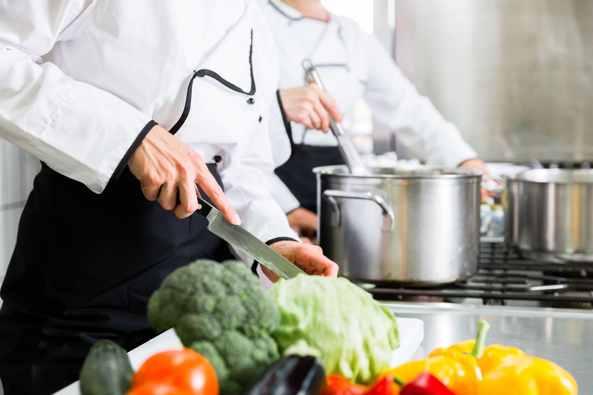 Köche in einem Betriebsrestaurant bereiten frisches Gemüse für eine gesunde Mahlzeit zu. Im Vordergrund liegt buntes, frisches Gemüse wie Brokkoli, Paprika und Tomaten, während im Hintergrund in großen Töpfen gekocht wird.