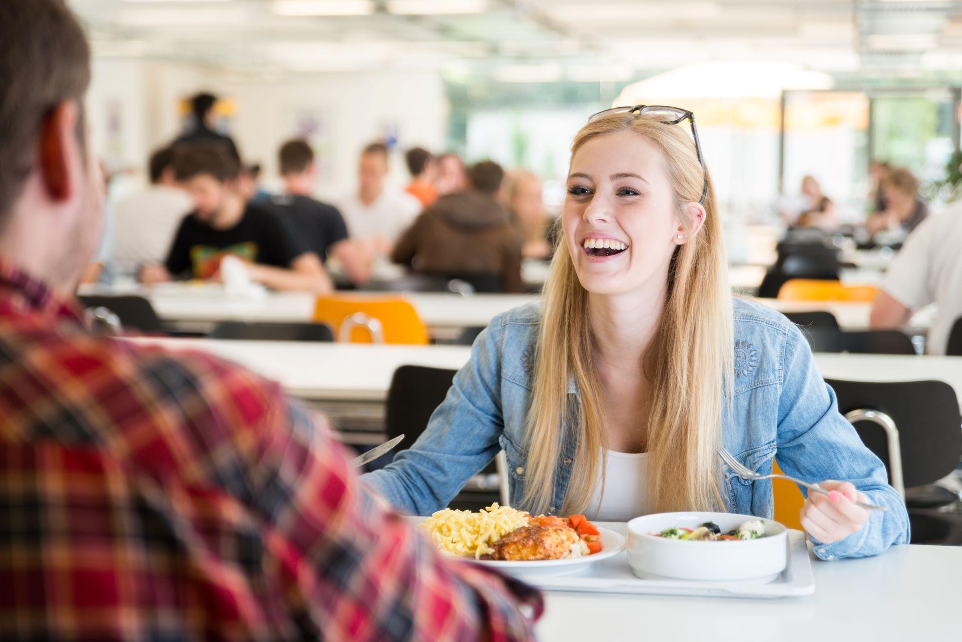 Eine junge Frau lacht während sie in einem gut besuchten Betriebsrestaurant mit einem Kollegen isst. Vor ihr steht ein Tablett mit einer ausgewogenen Mahlzeit, bestehend aus verschiedenen Gerichten. Die entspannte Atmosphäre und die Gemeinschaft in der Kantine tragen zum Wohlbefinden bei.