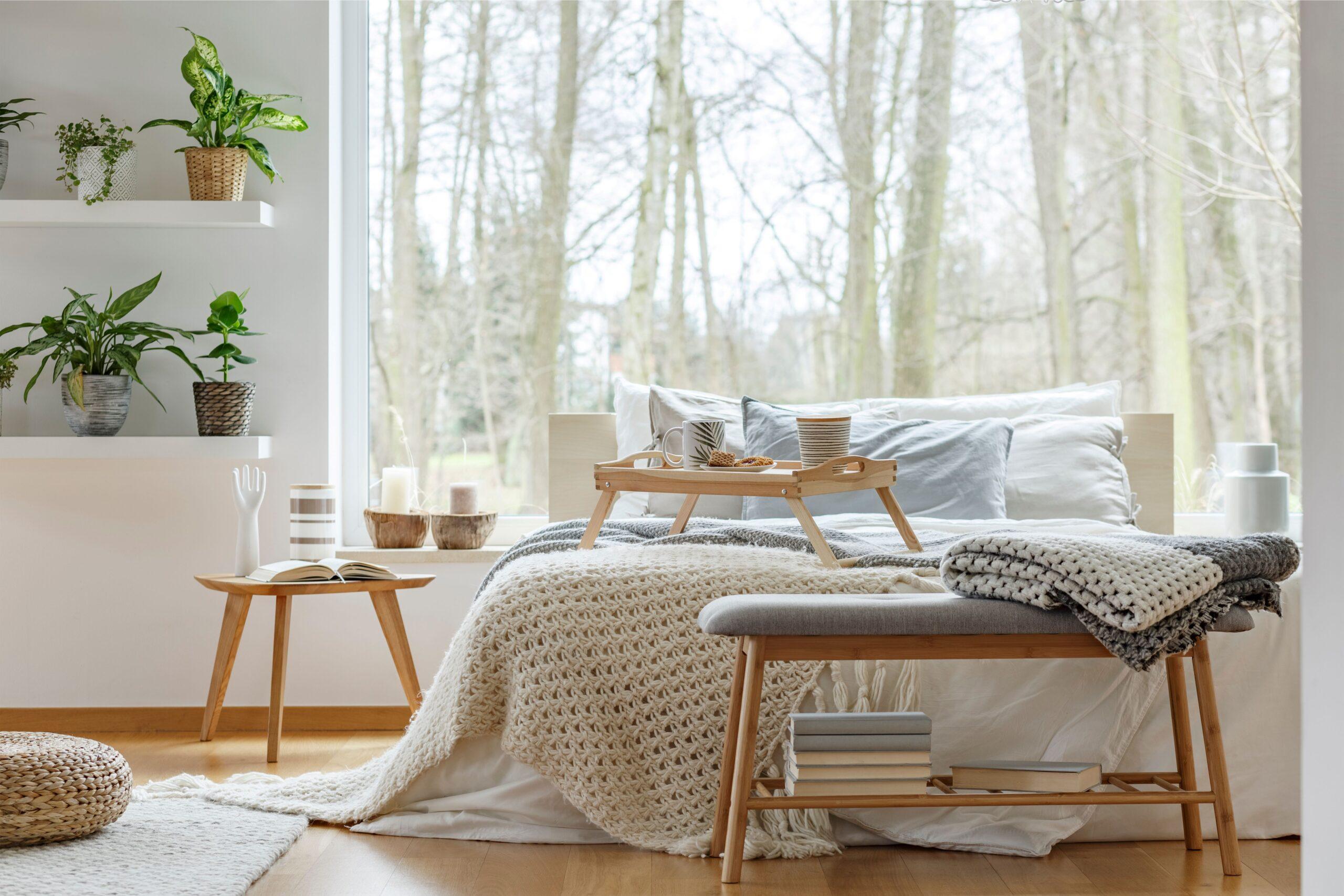 Helles Schlafzimmer mit großen Fenstern, einem gemütlich dekorierten Bett, und einer Holzbank ohne Lehne am Fußende des Bettes, umgeben von Pflanzen und natürlicher Dekoration.