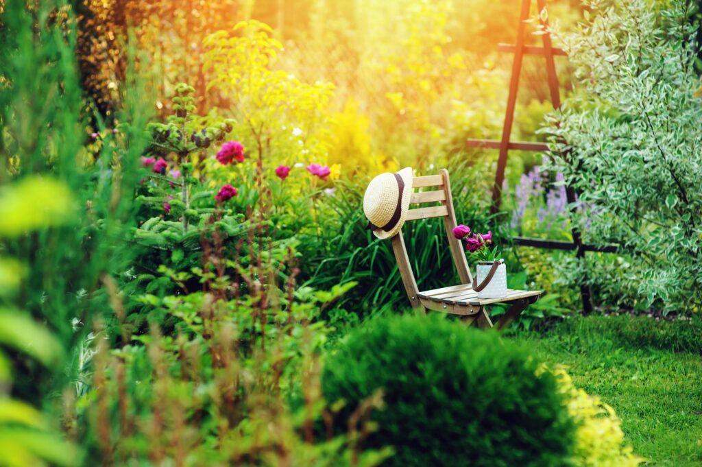 Ein Stuhl steht in einem Garten mit einer Blume und einem Hut drauf. Im Hintergrund scheint die Sonne durch das Gebüsch