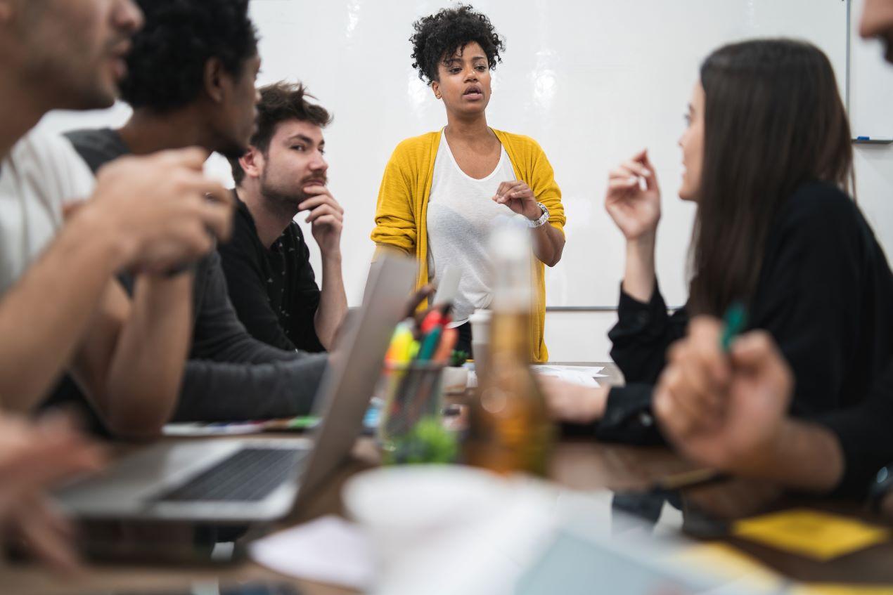 Eine junge Frau in einem gelben Cardigan leitet ein Teammeeting in einer Employer Branding Agentur, bei dem kreative Strategien zur Mitarbeitergewinnung besprochen werden.