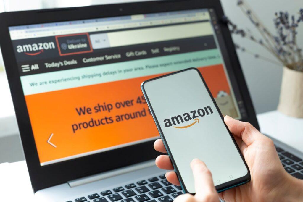 Person hält Smartphone mit Amazon-Logo vor Laptop mit Amazon-Website – Strategien für mehr Erfolg durch Amazon Agentur